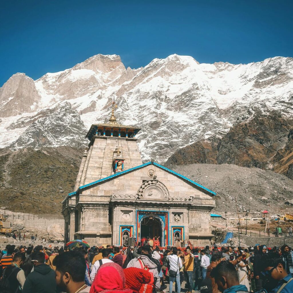 A crowd of pilgrims at Kedarnath Temple with snowcapped Himalayan peaks in the background.
